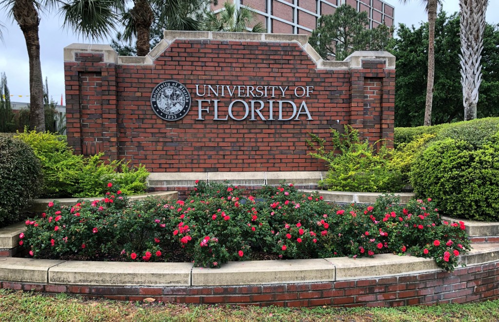 A brick sign displaying the University of Florida logo, surrounded by greenery and flowers under a clear sky.