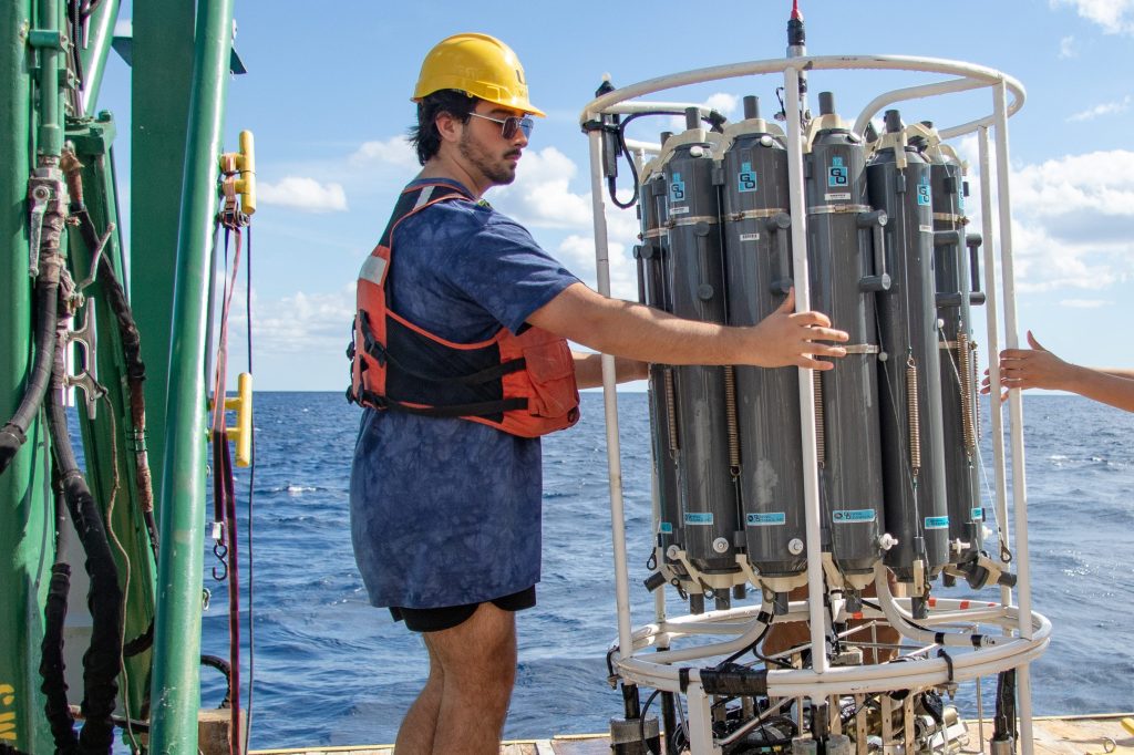 A man wearing a yellow helmet and a life jacket is standing on a dock, holding a large device, with water and a ship in the background under an open sky.