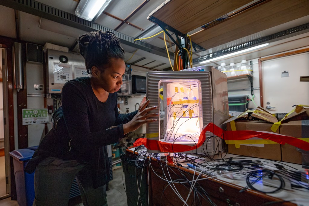 A woman is standing, examining a piece of equipment. The background features a wall and ceiling.