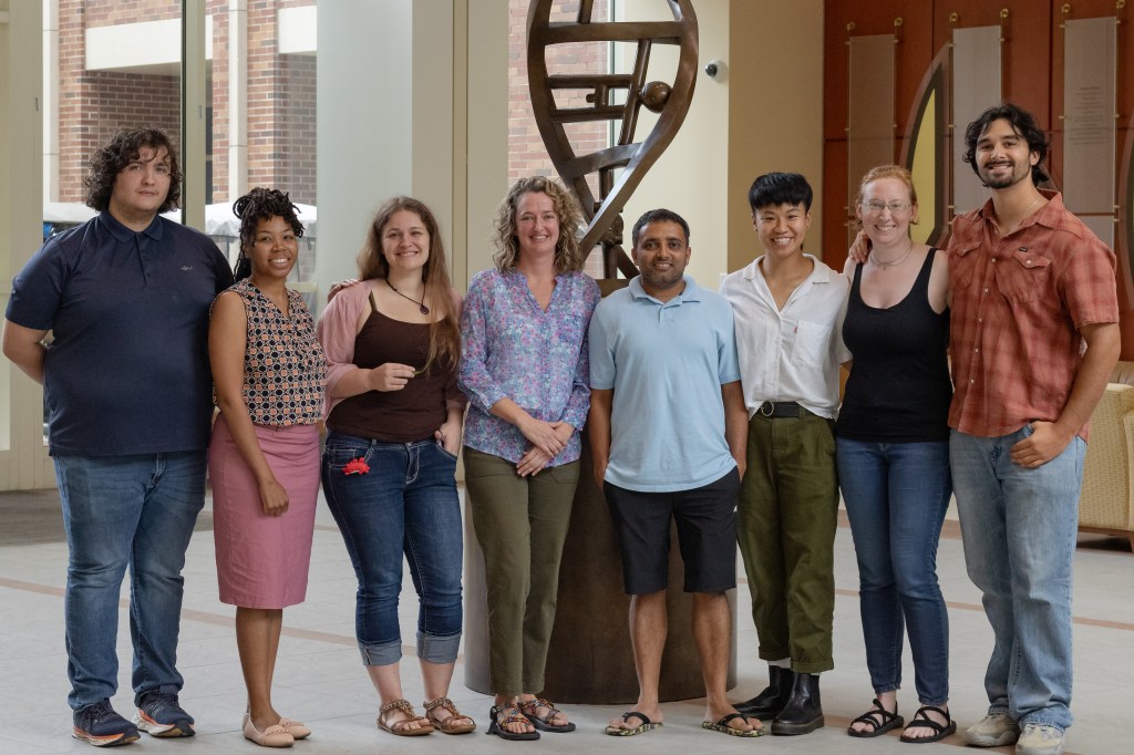 A group of people standing together indoors, smiling for a photo in front of a wall. They are dressed casually in jeans and various styles of footwear.