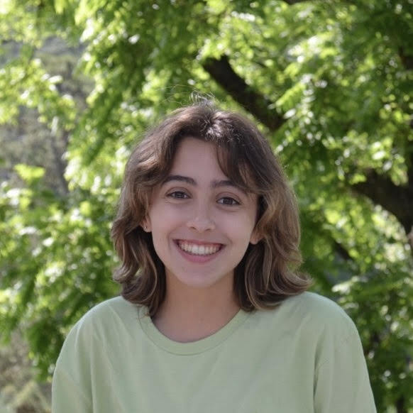 A young person with short, wavy hair smiles brightly while standing outdoors. They're wearing a light green shirt with lush green trees in the background.