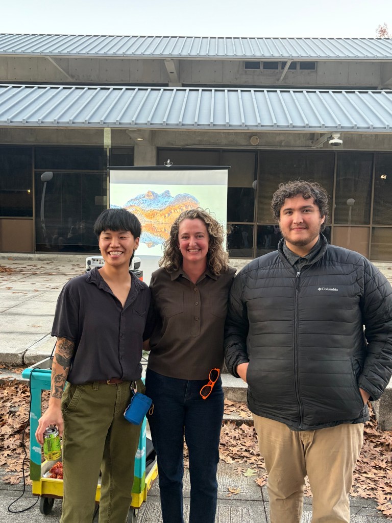 Three people smile at the camera while standing together outdoors. They are casually dressed with a background of a modern building. The mood is cheerful.