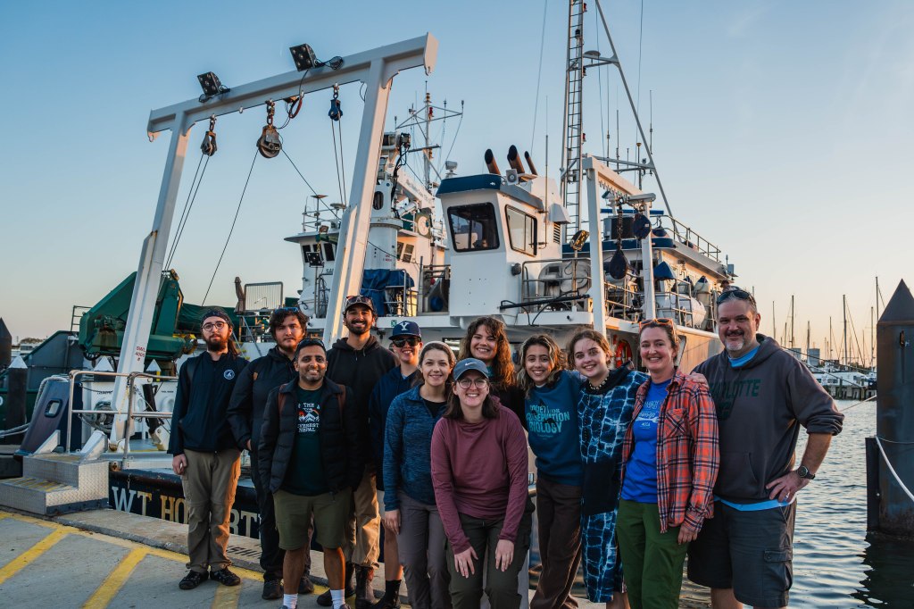 A group of people pose in front a researcher ship. The backdrop is sunset over the water.