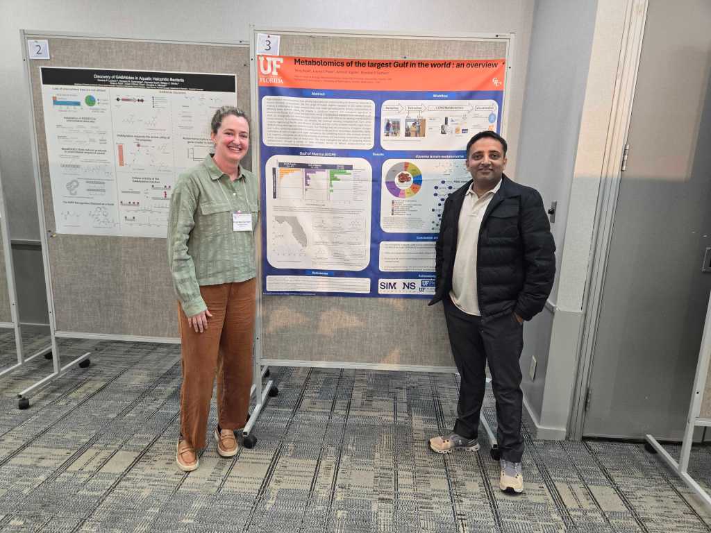 Two people smile at the camera while standing in front of a scientific poster. The mood is cheerful.