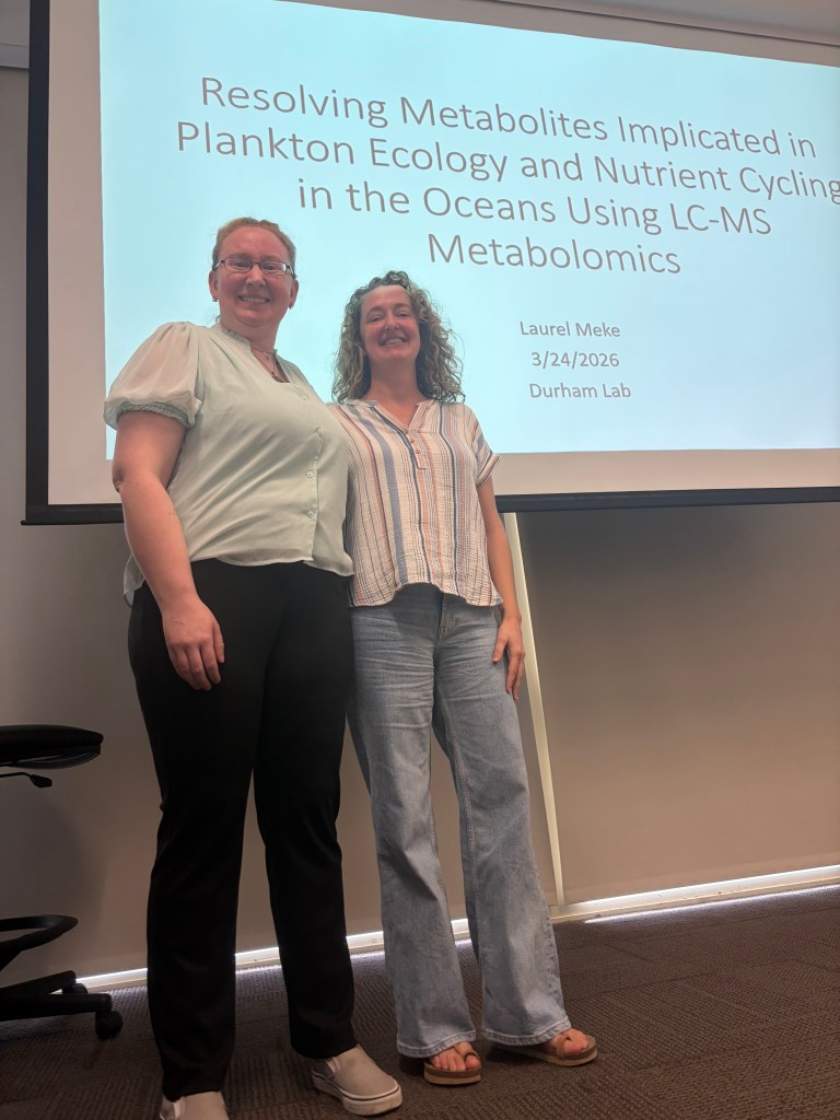Two smiling individuals stand in front of a presentation slide about ocean nutrient cycling and LC-MS metabolomics. The setting is casual and academic.