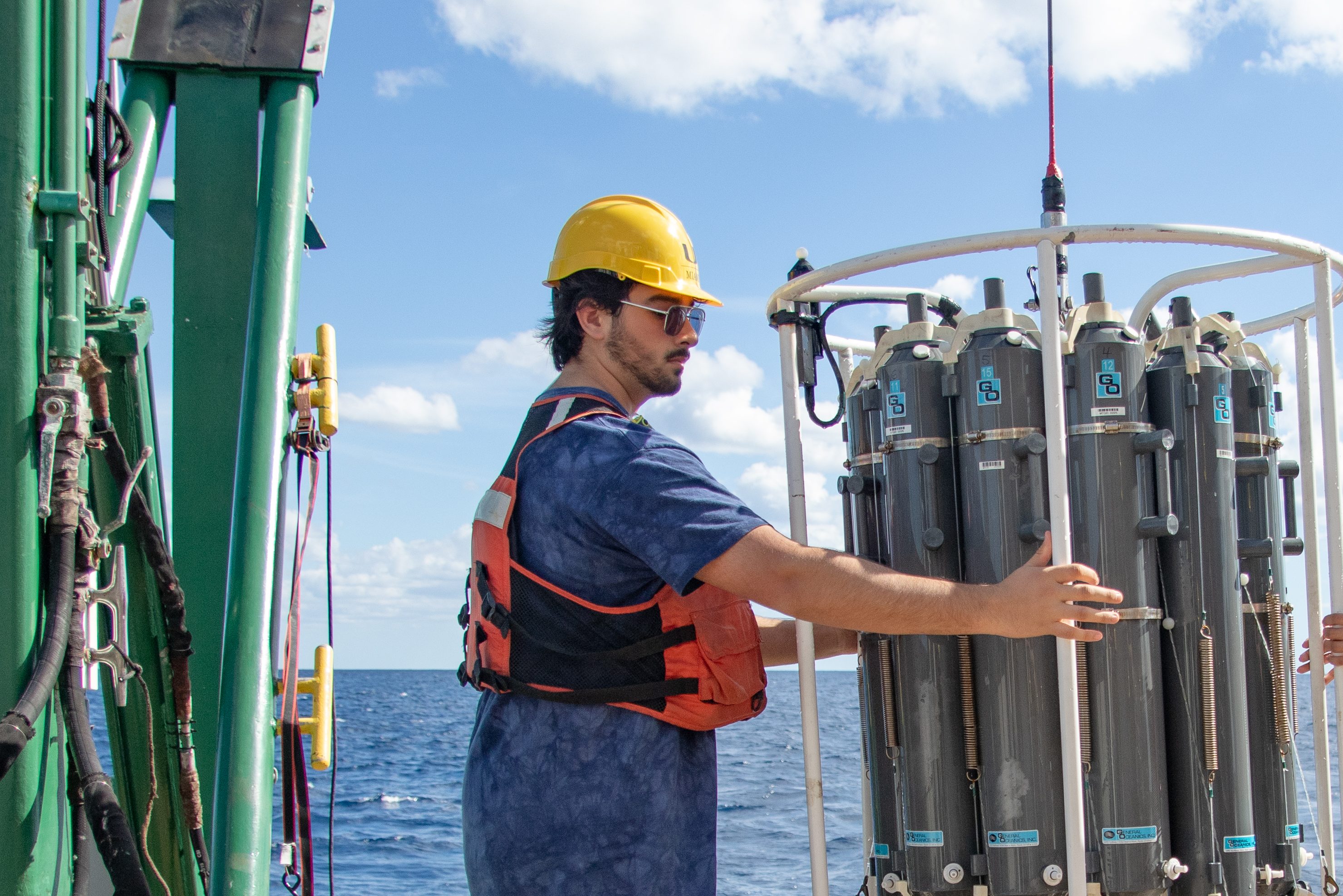 A person in safety gear handles oceanographic equipment on a ship's deck, with the ocean and blue sky in the background.