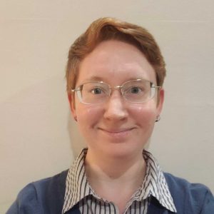 A person with short, reddish hair and glasses smiles warmly. They wear a blue sweater over a striped shirt. The background is plain and light-colored.