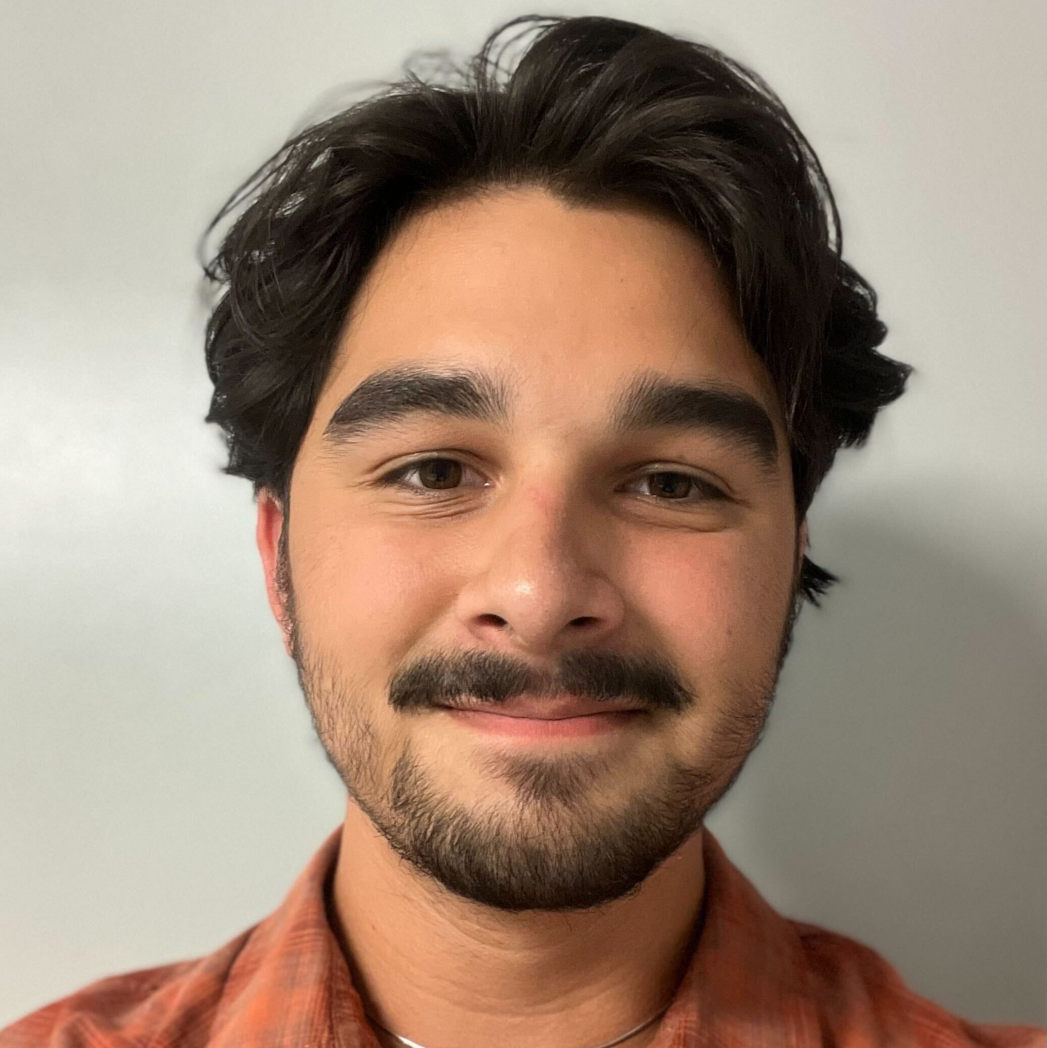 Young man with short, dark hair and a beard smiles warmly. He wears a red plaid shirt against a plain background, conveying a friendly, relaxed tone.