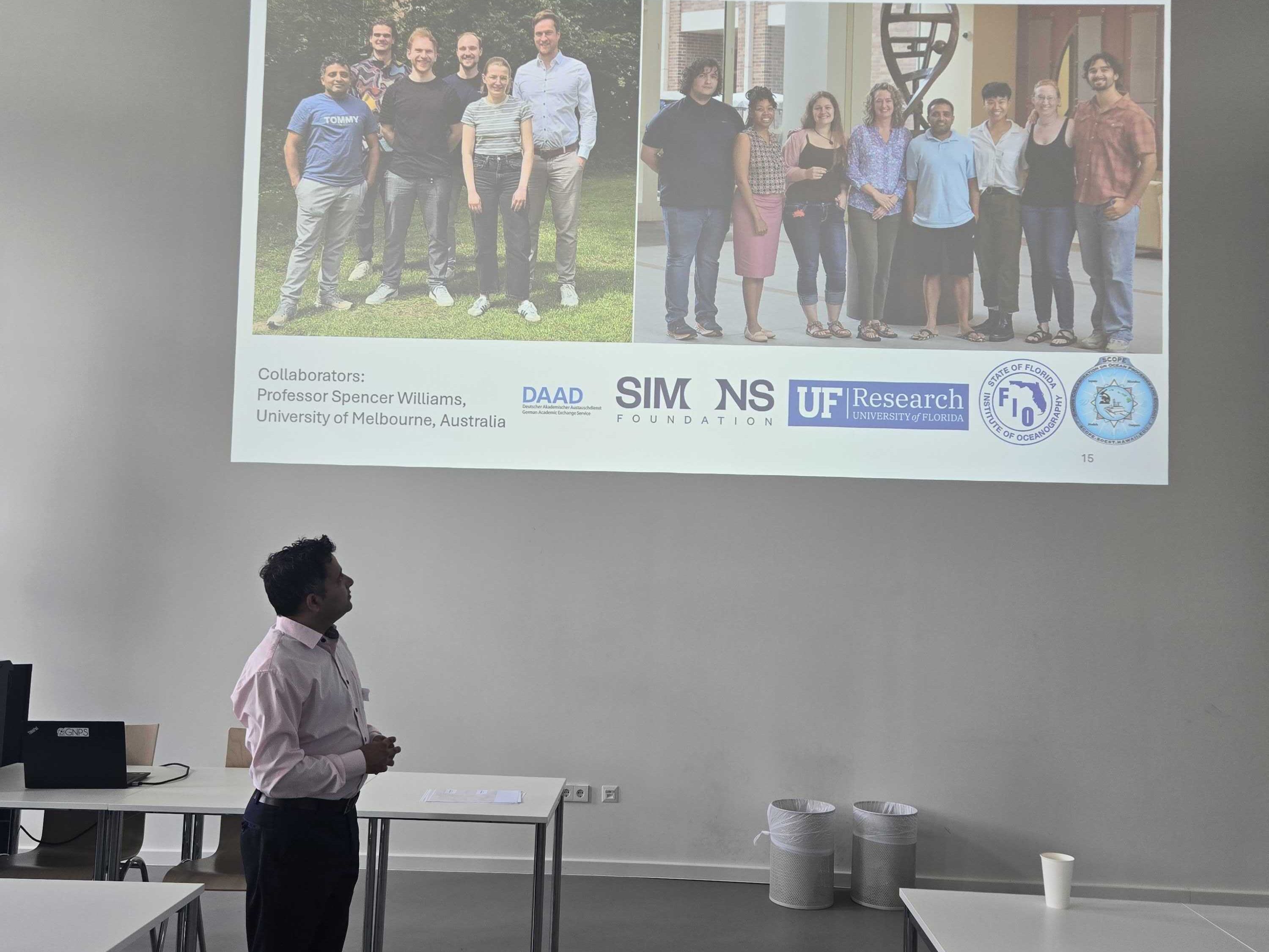 A person stands in a classroom presenting a slide with a group photo, logos, and a “Thank you!” message projected on the wall.