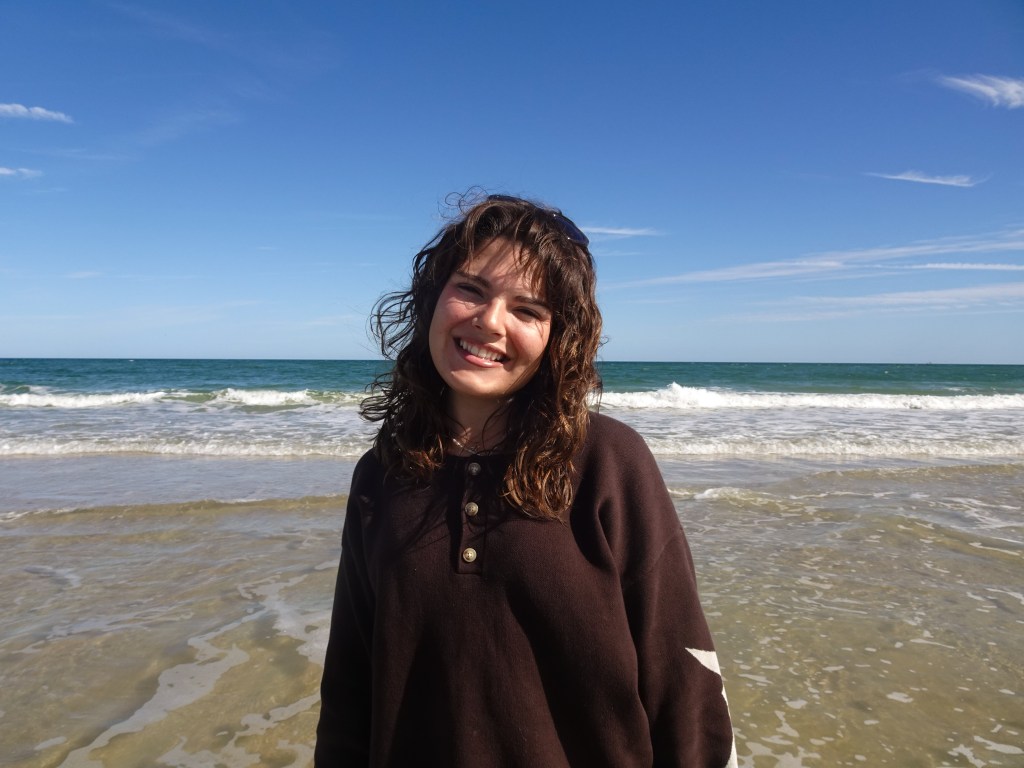 A woman with curly hair smiles at the beach, wearing a dark brown shirt, with the ocean and blue sky in the background.